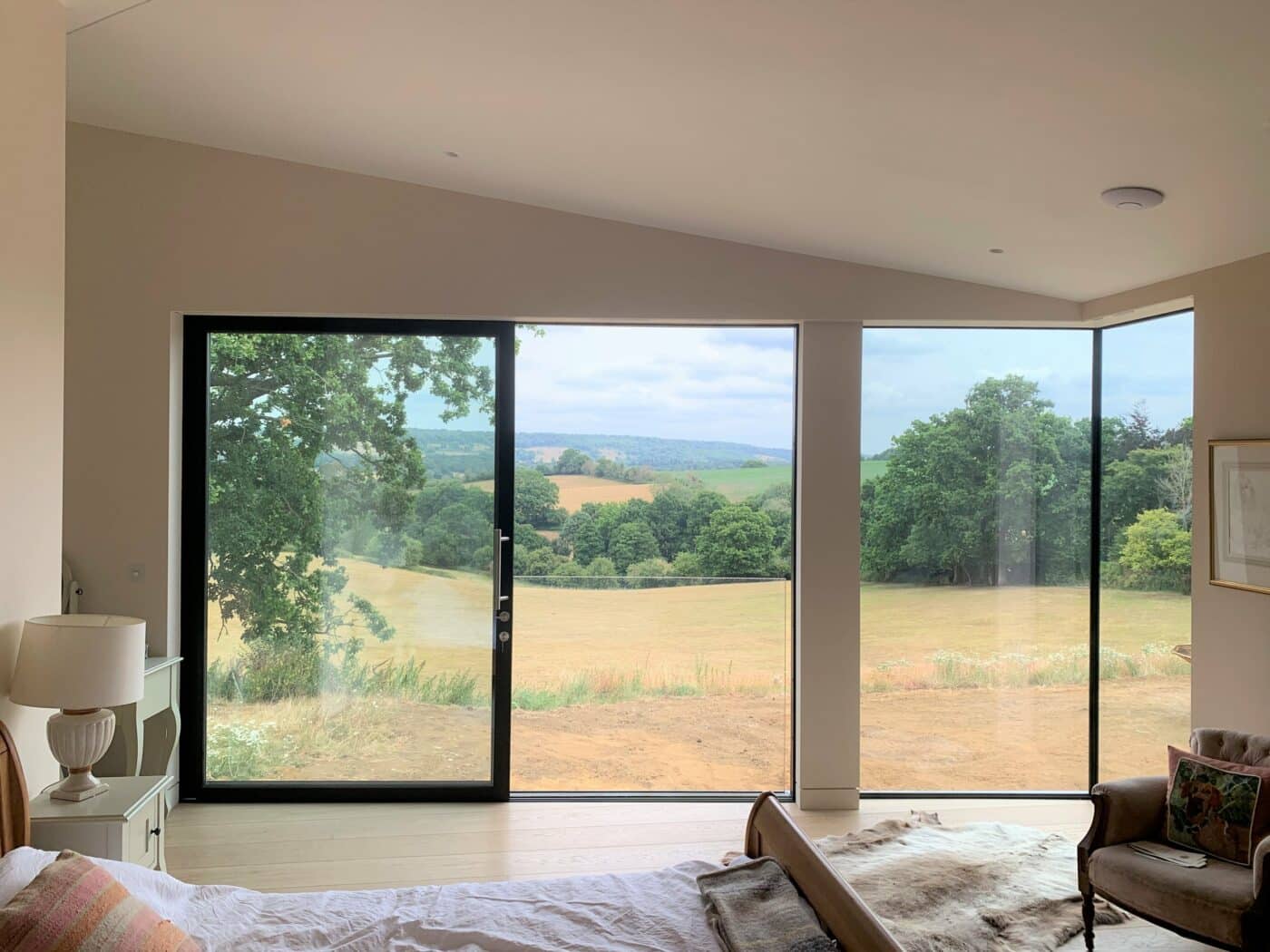 Modern bedroom with large glass windows and sliding door, offering a clear view of green fields, trees, and distant hills under a cloudy sky.