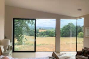 Modern bedroom with large glass windows and sliding door, offering a clear view of green fields, trees, and distant hills under a cloudy sky.