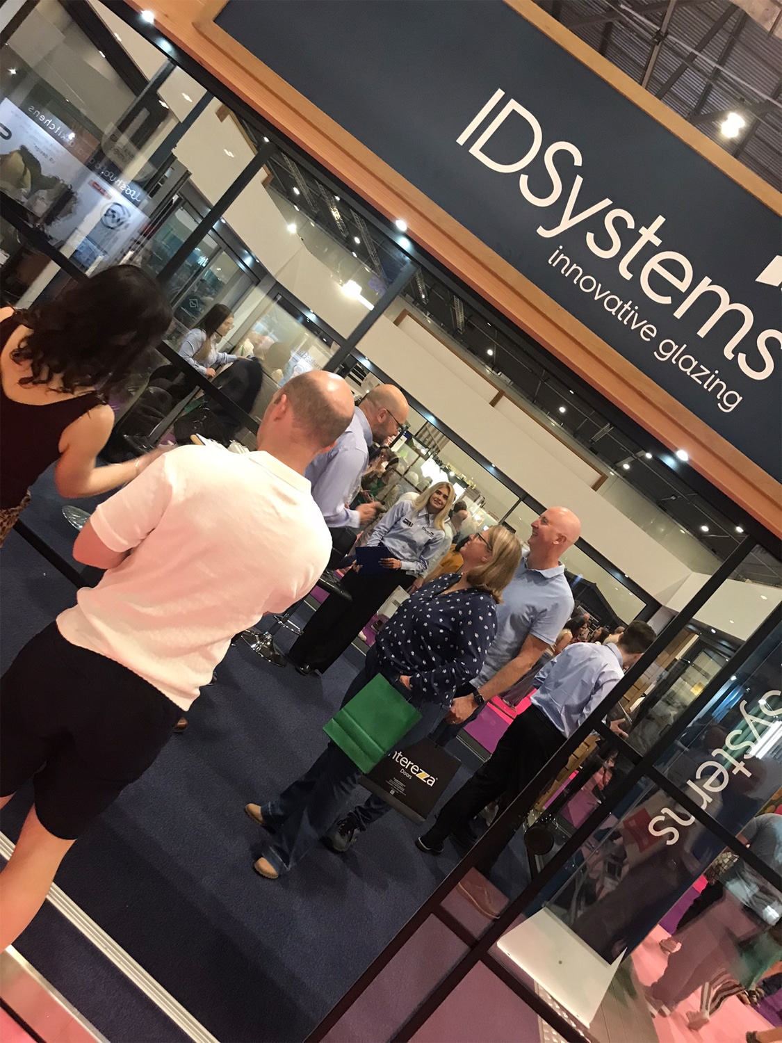 People stand and converse at the entrance of the IDSystems booth, which showcases innovative glazing products at Grand Designs Live