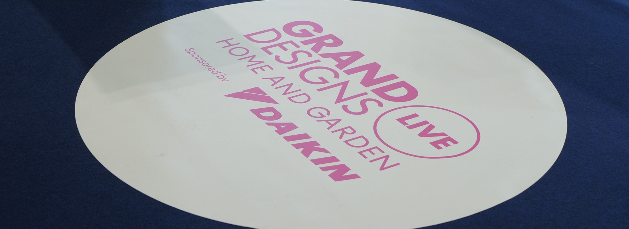 Grand Designs Live exhibition branding displayed on white circular signage, showcasing IDSystems' bespoke glazing solutions and aluminium door innovations.