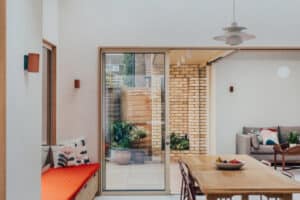 A modern dining area with a wooden table and chairs, built-in bench with red cushion, large sliding glass door, and a view of a patio and living room.