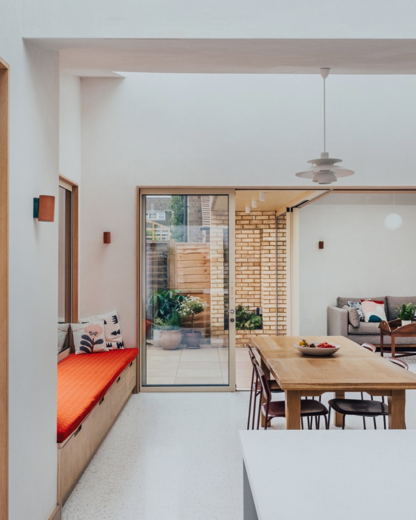 A modern dining area with a wooden table and chairs, built-in bench with red cushion, large sliding glass door, and a view of a patio and living room.