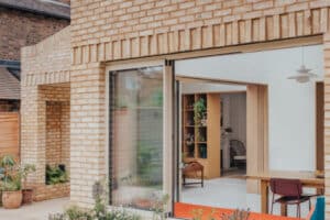 A modern brick house with large sliding glass doors opens to a patio with a raised flower bed and potted plants. Indoor seating and shelves are visible through the doorway.