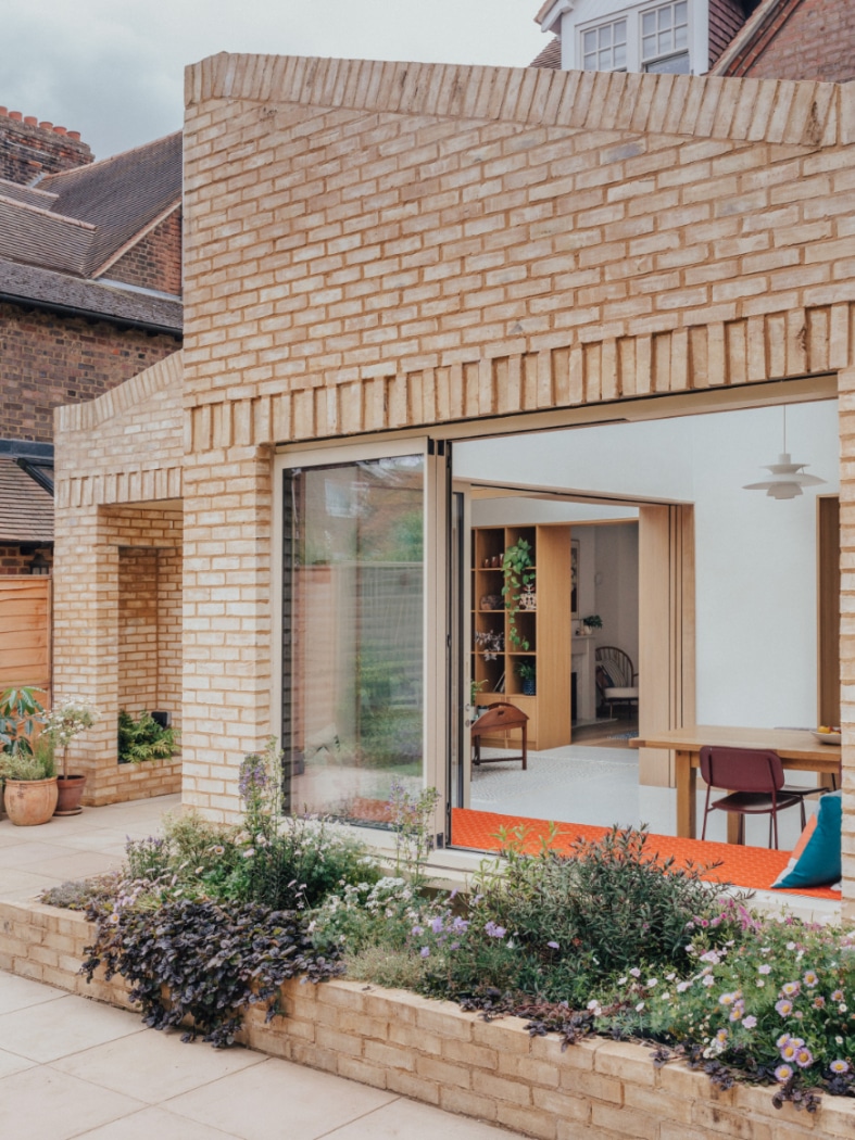 A modern brick house with large sliding glass doors opens to a patio with a raised flower bed and potted plants. Indoor seating and shelves are visible through the doorway.