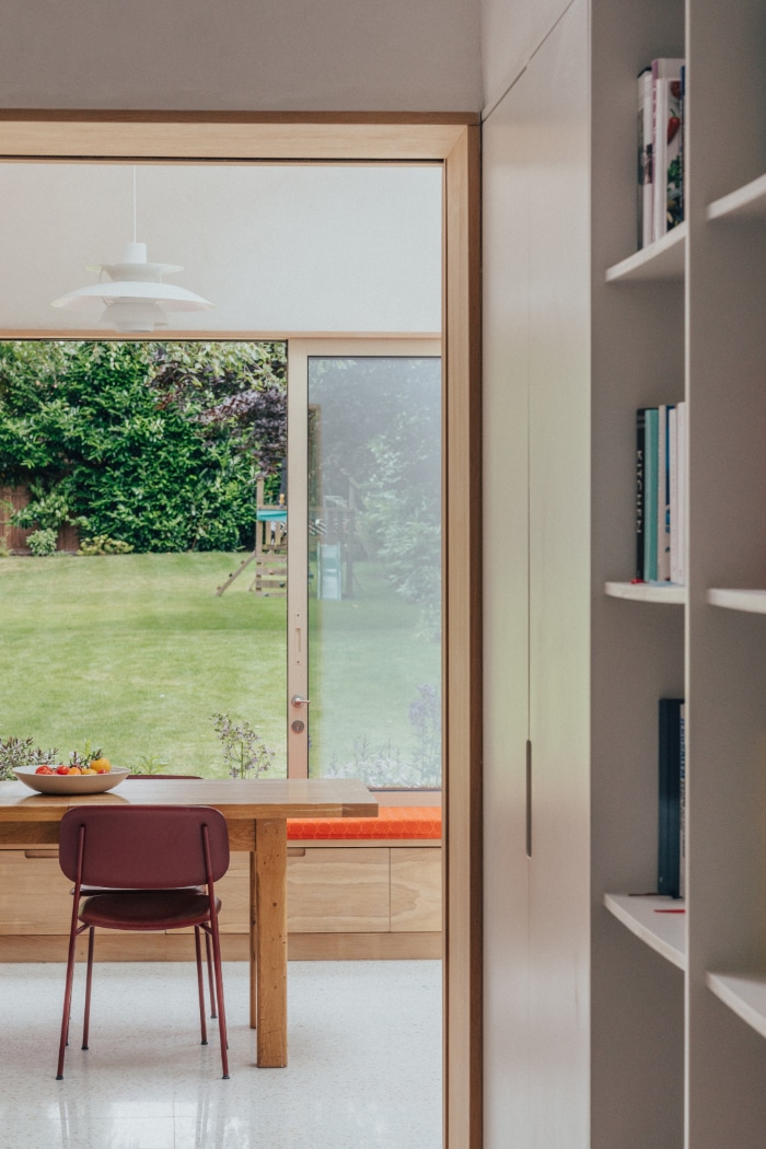 A dining area with a wooden table and red chair overlooks a green backyard through large glass doors; bookshelf is visible in the foreground.