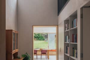 A hallway with a patterned rug, bookshelves, a wooden cabinet, houseplants, and a view into a dining area with large windows overlooking a green outdoor space.