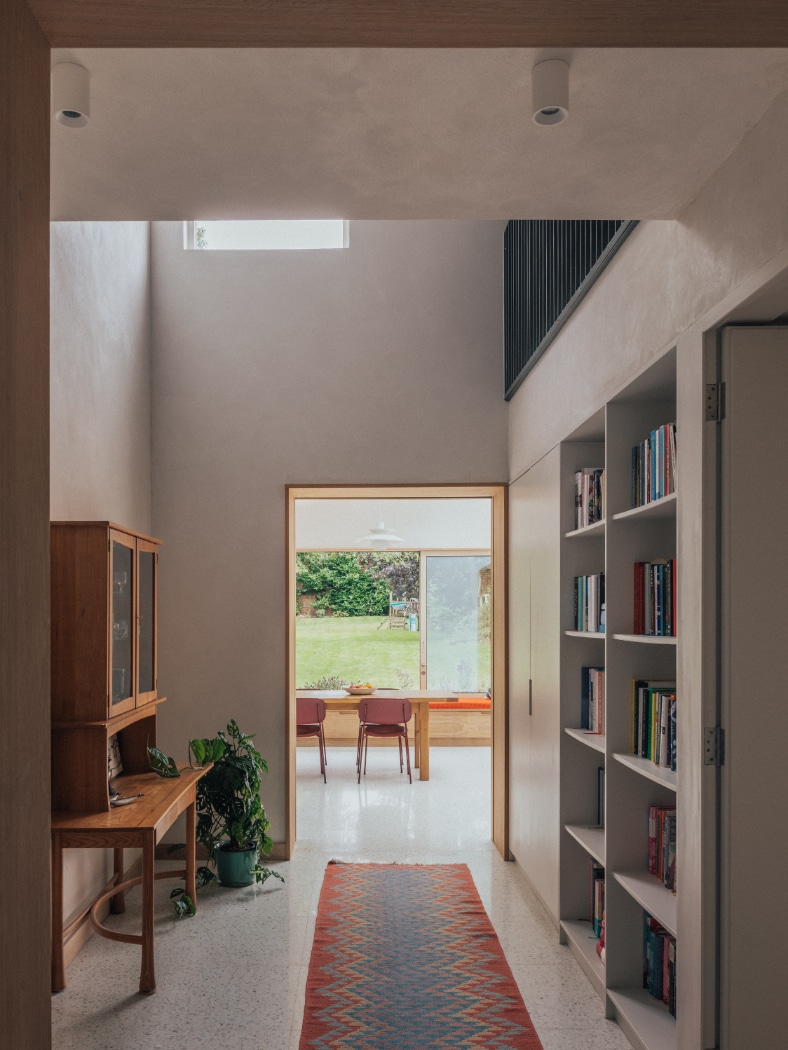 A hallway with a patterned rug, bookshelves, a wooden cabinet, houseplants, and a view into a dining area with large windows overlooking a green outdoor space.