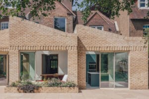 Modern brick house extension with large glass sliding doors, zigzag roof design, and a patio overlooking a green lawn. Original older brick house visible in the background.