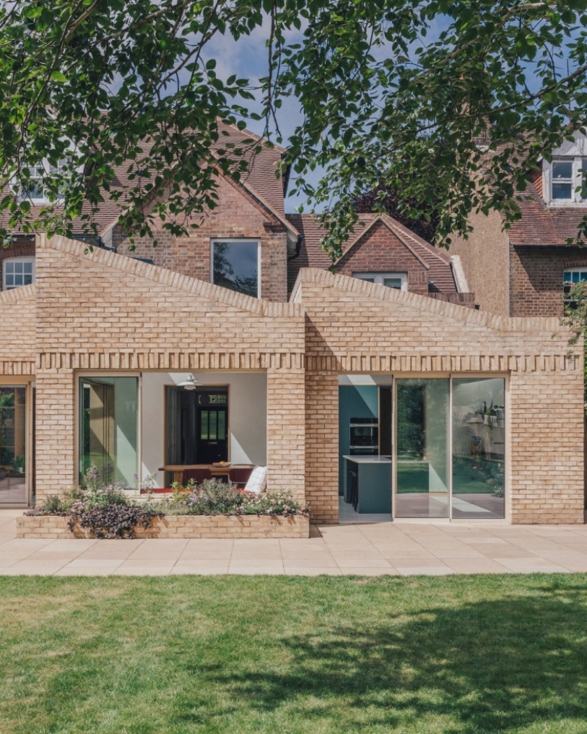 Modern brick house extension with large glass sliding doors, zigzag roof design, and a patio overlooking a green lawn. Original older brick house visible in the background.