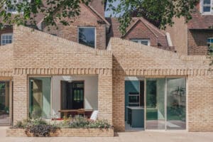 Modern brick extension with zigzag roofline, large glass doors, and windows, attached to an older brick house with a garden visible inside.