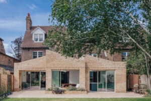 A modern brick extension with large glass doors is attached to the rear of a traditional two-story house, with a tree and garden in the foreground.