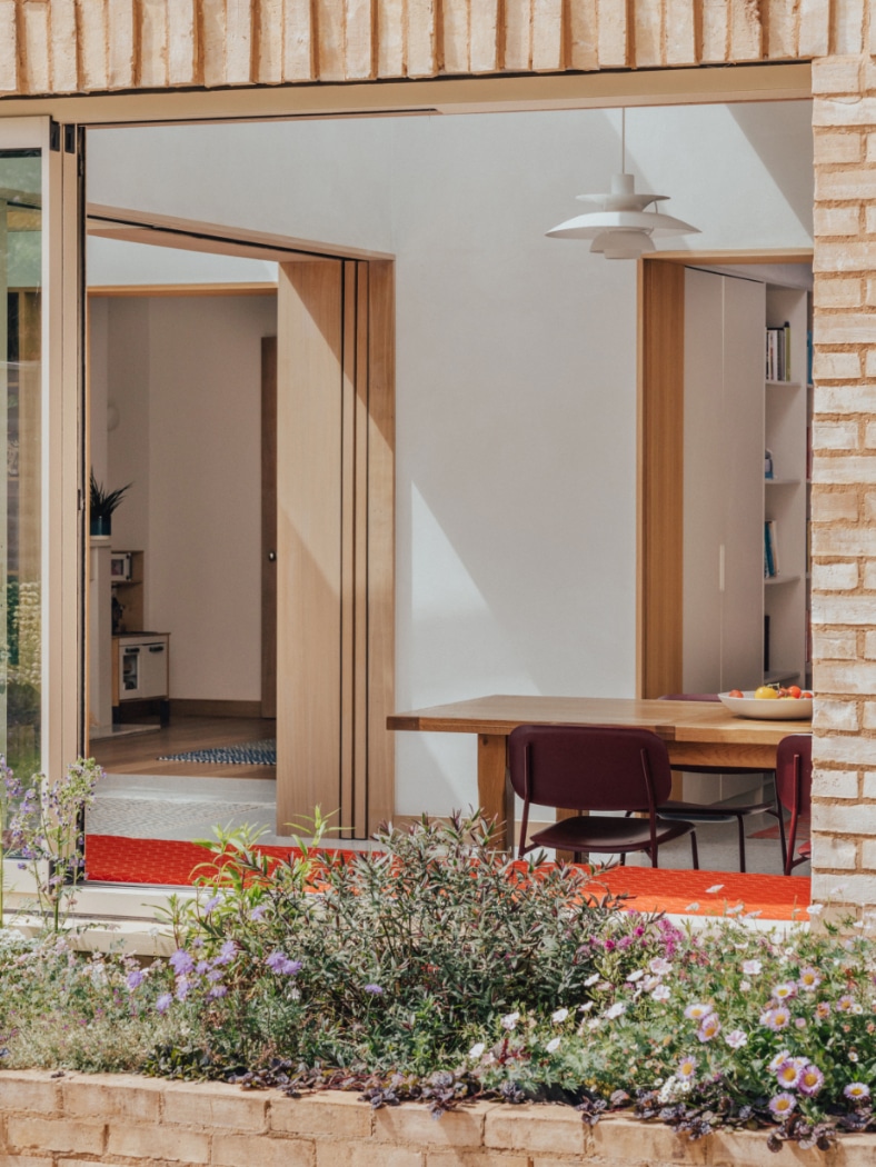 A modern interior with light wood furniture, red chairs, and a long table is visible through a large open window, with flowering plants and shrubs in the foreground.