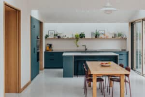 Modern kitchen with blue cabinets, an island, and a wooden dining table with six chairs. Shelves with plants and dishes are mounted on the wall above the countertop.