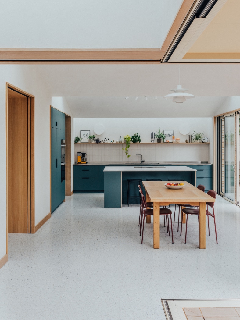 Modern kitchen with blue cabinets, an island, and a wooden dining table with six chairs. Shelves with plants and dishes are mounted on the wall above the countertop.