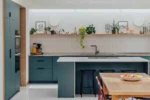 Modern kitchen with teal cabinets, a central island with bar stools, open shelving with plants, and a wooden dining table with fruit bowl and red chairs.