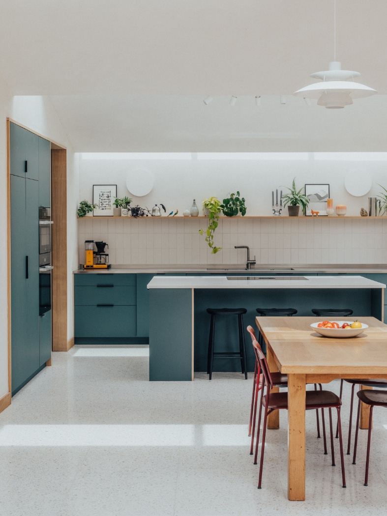 Modern kitchen with teal cabinets, a central island with bar stools, open shelving with plants, and a wooden dining table with fruit bowl and red chairs.
