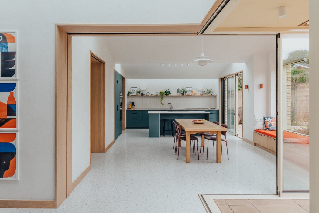 Contemporary open-plan kitchen and dining area with blue cabinetry, wooden table, chairs, and garden view through large sliding glass doors.