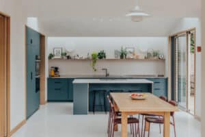 Modern kitchen with teal cabinets, a large island, open shelves with plants and decor, wooden dining table with red chairs, and sliding glass doors letting in natural light.