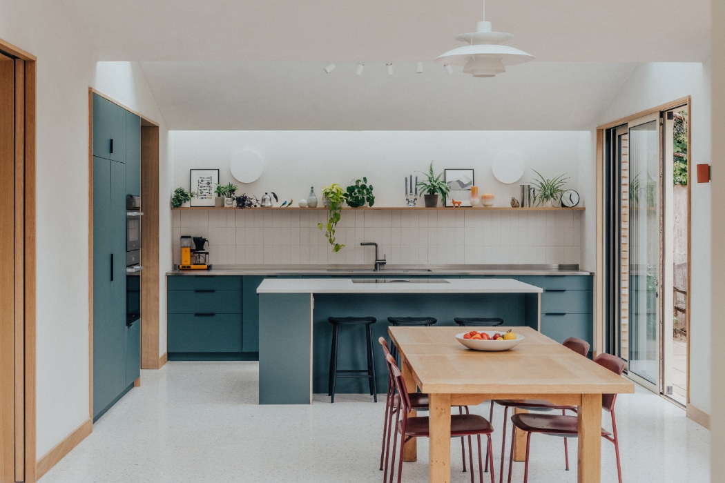 Modern kitchen with teal cabinets, a large island, open shelves with plants and decor, wooden dining table with red chairs, and sliding glass doors letting in natural light.