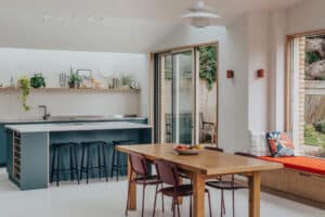 Modern kitchen and dining area with a wooden table, four chairs, a built-in bench with cushions, and a minimalist island with black stools, next to large glass doors opening to a garden.