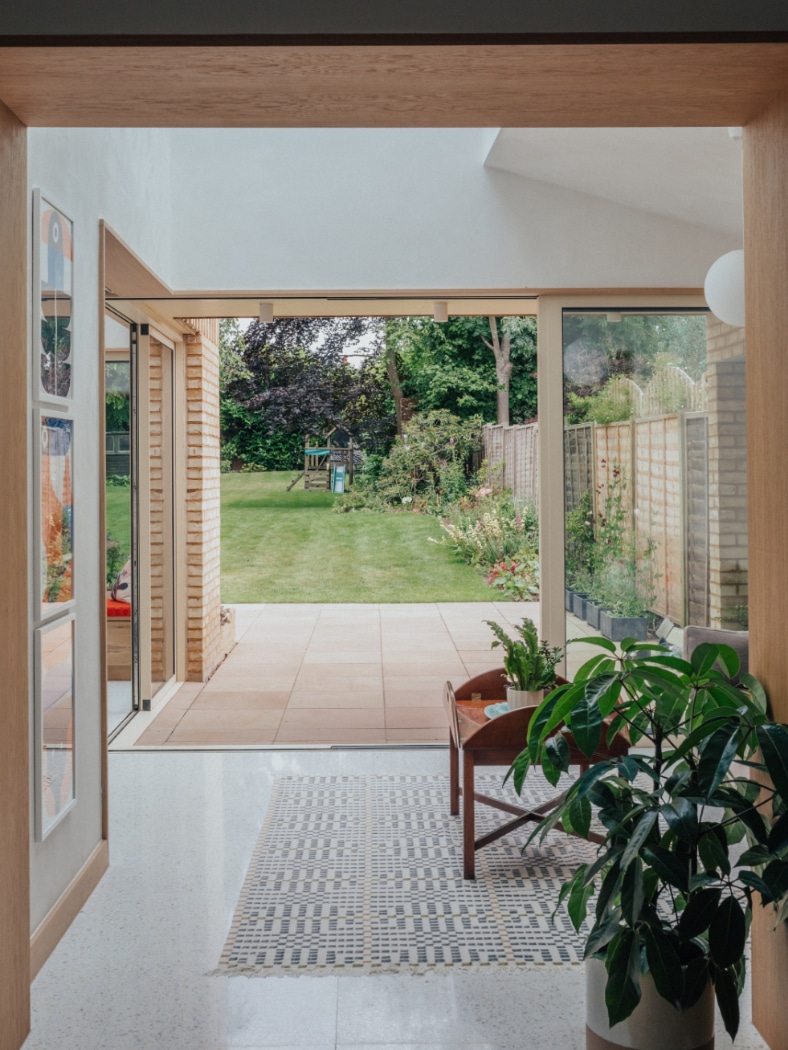 A modern living space with large glass doors opens onto a patio and a green backyard with a children’s playset in the distance. Indoor plants and a patterned rug are in the foreground.