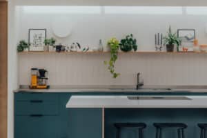 Modern kitchen with teal cabinets, a white countertop island with two black stools, a yellow coffee machine, and a shelf displaying plants and decor against a white tiled wall.