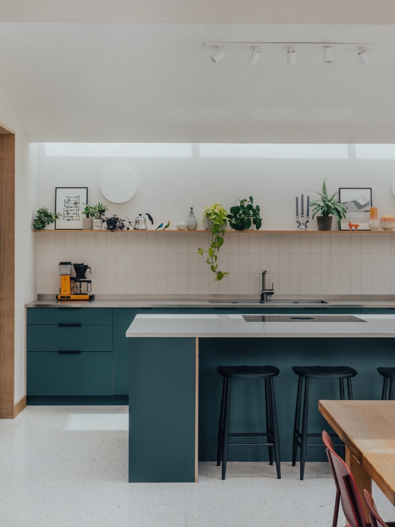 Modern kitchen with teal cabinets, a white countertop island with two black stools, a yellow coffee machine, and a shelf displaying plants and decor against a white tiled wall.
