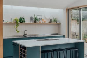 A modern kitchen with teal cabinets, a white island with black barstools, open shelving, plants, and glass doors leading to an outdoor area.