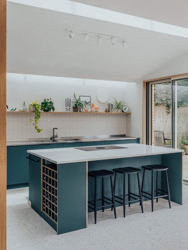 A modern kitchen with teal cabinets, a white island with black barstools, open shelving, plants, and glass doors leading to an outdoor area.