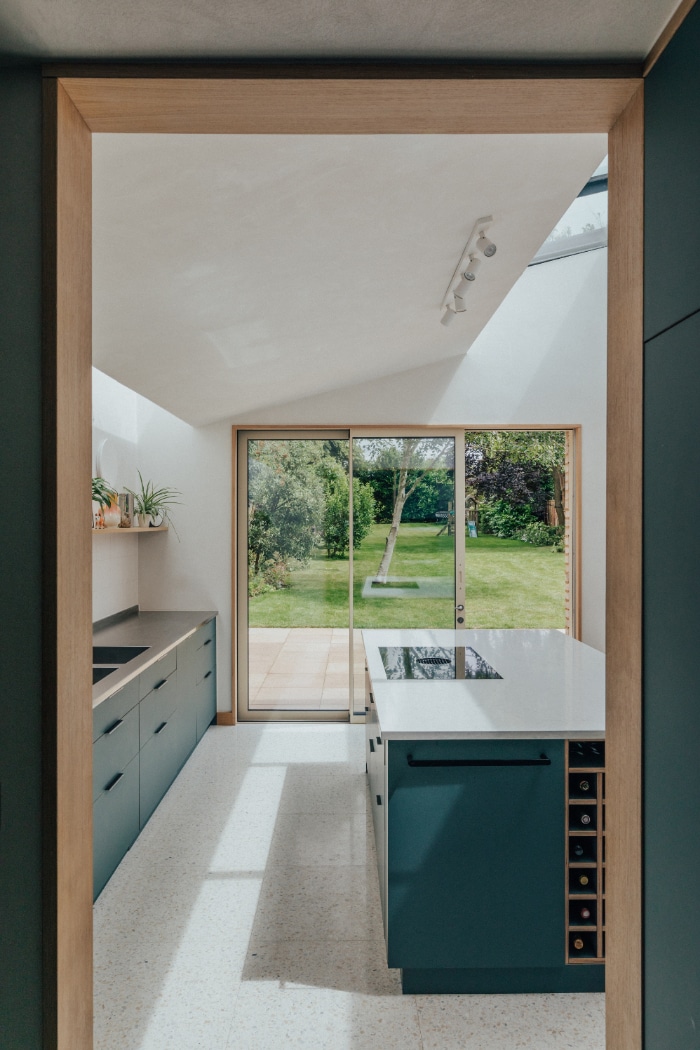 Modern kitchen with dark cabinetry, a central island, and large glass doors opening to a green backyard. Natural light fills the space through windows and a skylight.