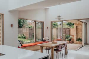 Modern dining area with a wooden table and four chairs, large windows, a built-in bench with a red cushion, and views of a garden and patio outside.