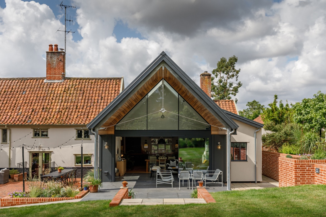 A modern home extension with large glass doors opens to a patio with outdoor seating, attached to a traditional house with a red-tiled roof and surrounded by grass and brickwork.