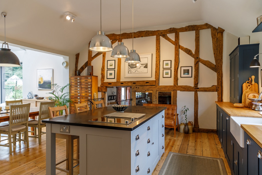 A rustic kitchen with exposed timber beams, a central island, modern pendant lights, dining table, and a mix of wooden and painted cabinetry on a wooden floor.