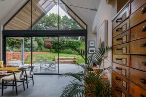 Modern living space with large glass doors opening to a patio and garden, featuring wooden furniture, potted plants, and a chest of drawers in the foreground.