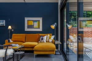 A modern living room with a mustard yellow sectional sofa, geometric artwork on a navy blue wall, a floor lamp, and large glass doors showing a patio with brick and outdoor seating.