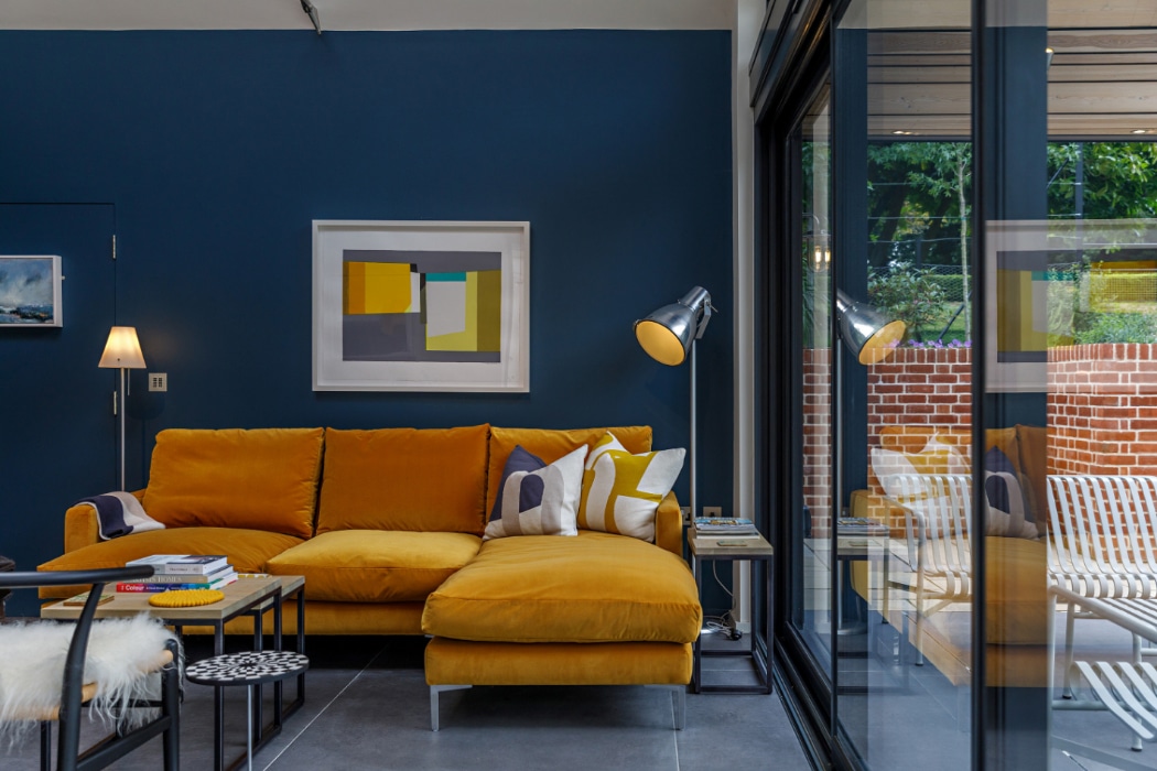 A modern living room with a mustard yellow sectional sofa, geometric artwork on a navy blue wall, a floor lamp, and large glass doors showing a patio with brick and outdoor seating.