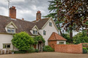 Large house with cream-colored walls, red-tiled roof, multiple chimneys, and a brick wall. Surrounded by trees and greenery, with a gravel driveway in front.