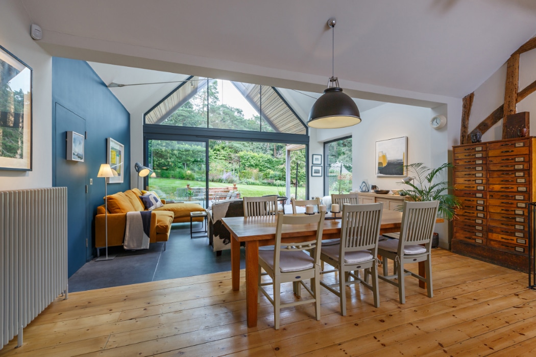 Open-plan dining and living area with wooden floors, modern furniture, large glass doors, and a view of a green garden outside.
