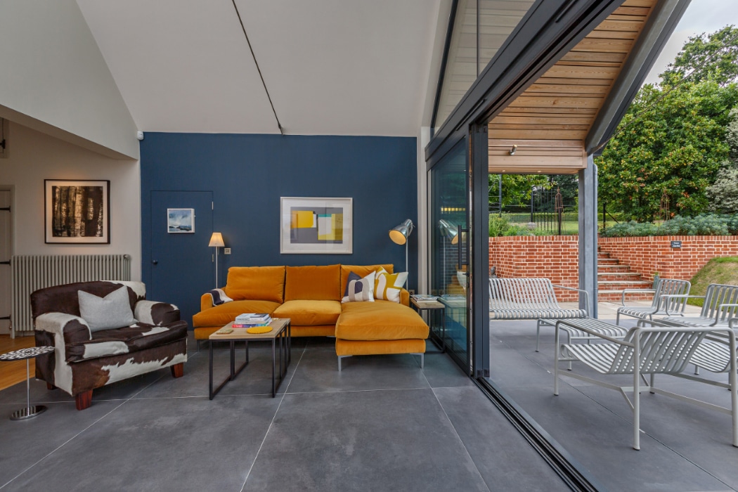 Modern living room with yellow sectional sofa, accent chair, and art on a blue wall; large glass doors open to an outdoor patio with metal chairs and a brick wall.