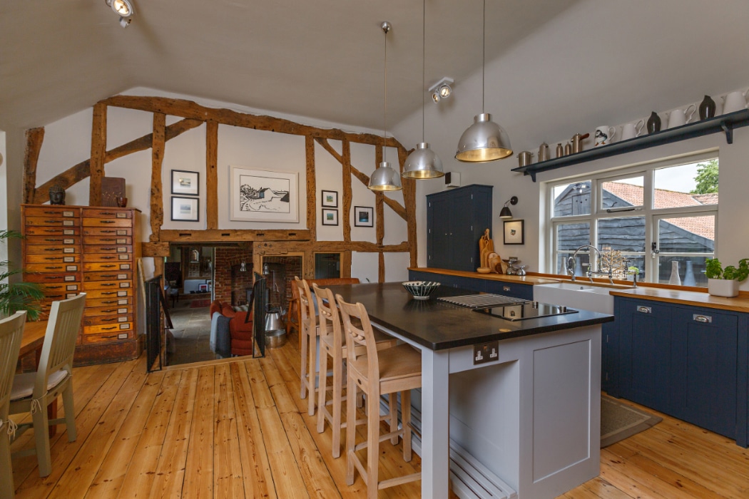A rustic kitchen with wooden beams, a large island with bar stools, pendant lights, blue cabinets, and a window above the sink. Dining table and vintage drawers are visible in the background.