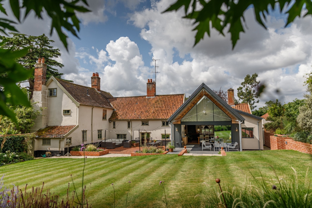 A large house with red-tiled roofs, multiple chimneys, and a modern glass extension overlooks a well-kept lawn and garden under a partly cloudy sky.