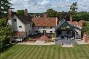 A large country house with red-tiled roofs, multiple chimneys, and a modern glass extension, surrounded by a lawn and patio area under a partly cloudy sky.