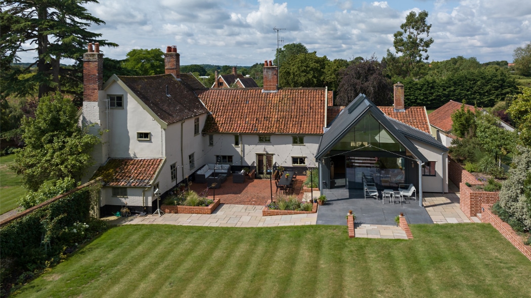 A large country house with red-tiled roofs, multiple chimneys, and a modern glass extension, surrounded by a lawn and patio area under a partly cloudy sky.