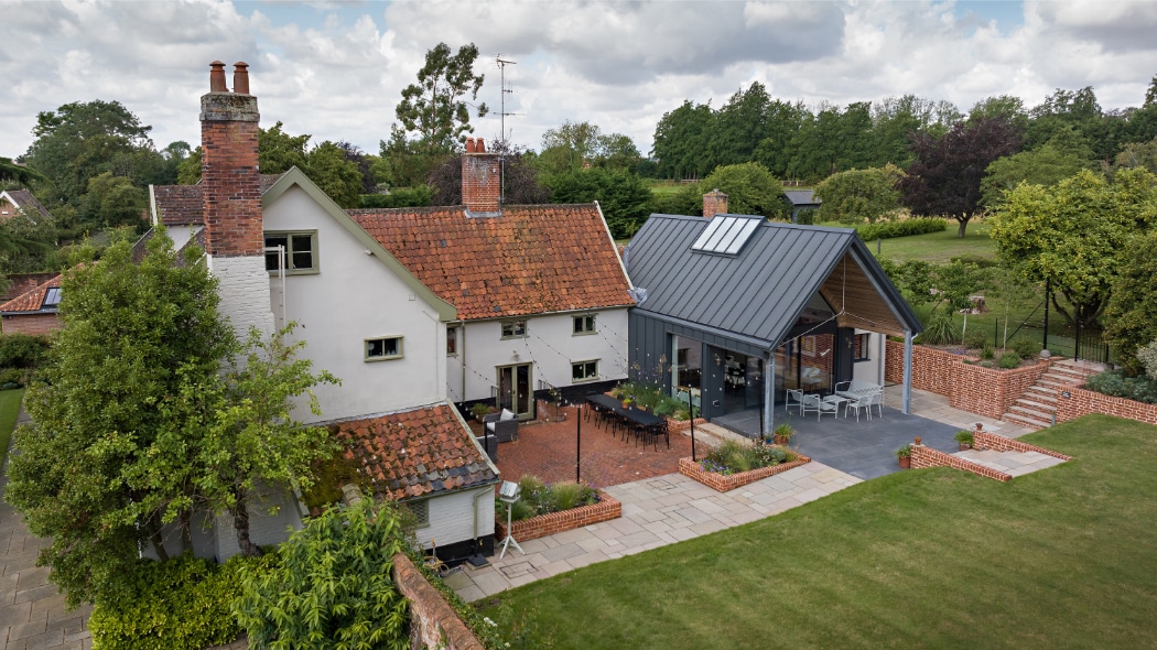 Aerial view of a traditional house with red-tiled roof, a modern extension with metal roofing, patio areas, and landscaped garden.