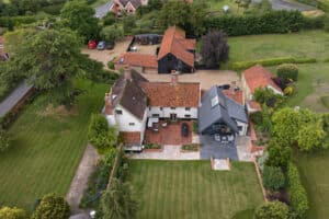 Aerial view of a large country house with red-tiled roofs, outbuildings, gardens, lawns, and a parking area surrounded by trees and greenery.