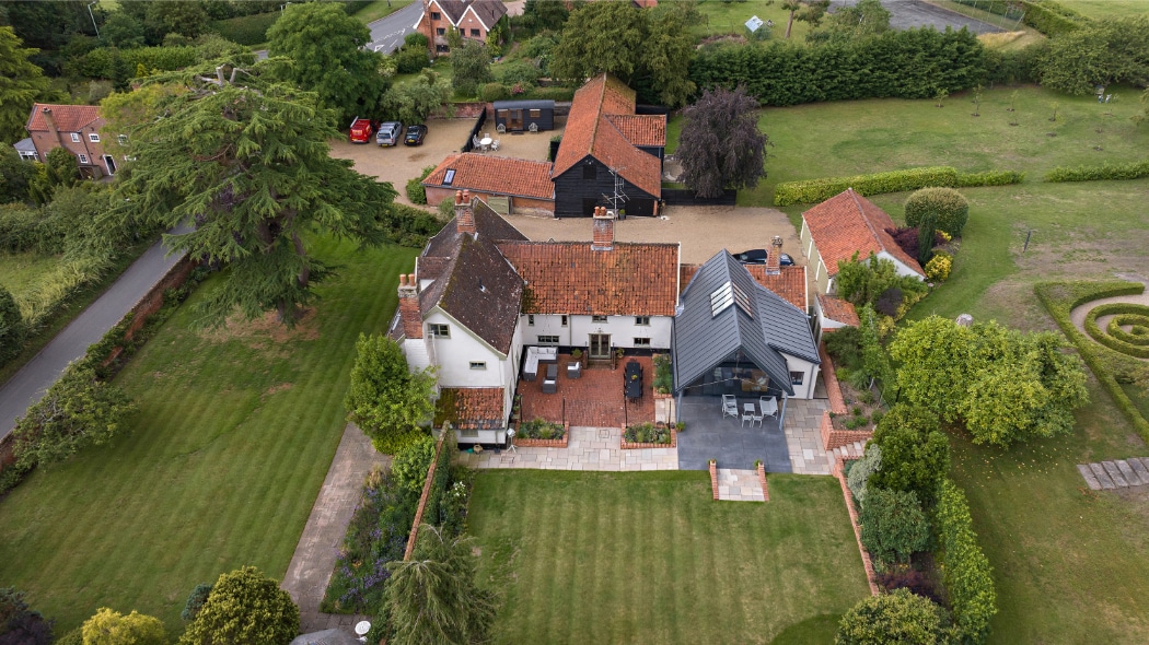 Aerial view of a large country house with red-tiled roofs, outbuildings, gardens, lawns, and a parking area surrounded by trees and greenery.