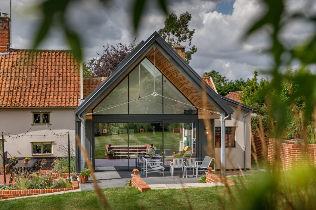 Modern house extension with large glass windows, outdoor seating area, and garden view, framed by leaves in the foreground.