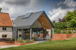 A modern house extension with a metal roof, large glass windows, and outdoor seating, connected to a traditional building with red tiles and surrounded by a landscaped garden.