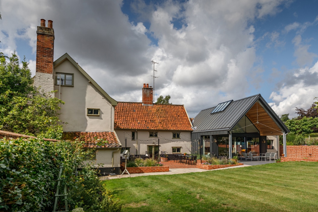 A traditional house with red tile roof and modern glass extension sits beside a lawn under a partly cloudy sky.