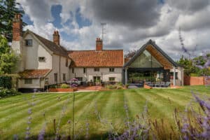 A traditional house with tiled roofs and brick chimneys features a modern glass extension, set on a well-kept lawn with cloudy skies overhead.